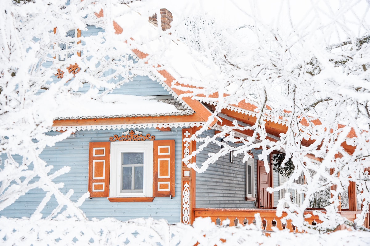 A charming cottage is enveloped in a thick layer of fresh snow, showcasing colorful orange trim against pale blue siding. Frosted branches frame the scene, accentuating the wintery ambiance. Intricate woodwork details add character to the cozy home.