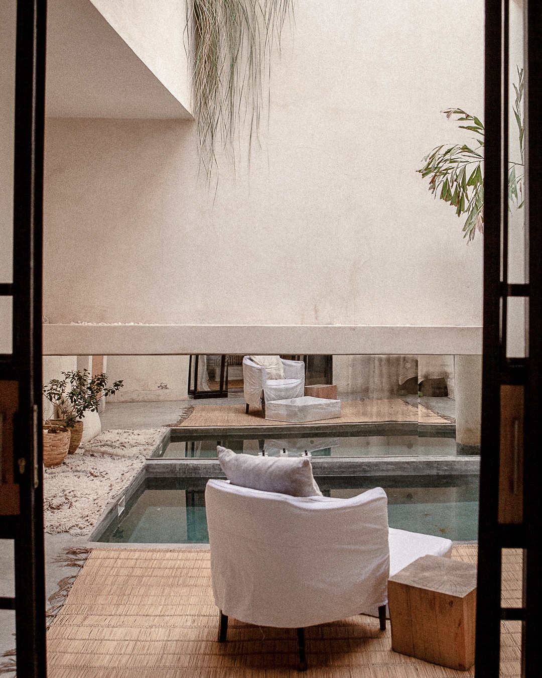 An inside view of the riad reveals a serene ambiance, featuring a small pool surrounded by minimalist furnishings. Two white chairs provide seating near the pool, while lush greenery is seen in the background, adding a touch of nature to the tranquil space.