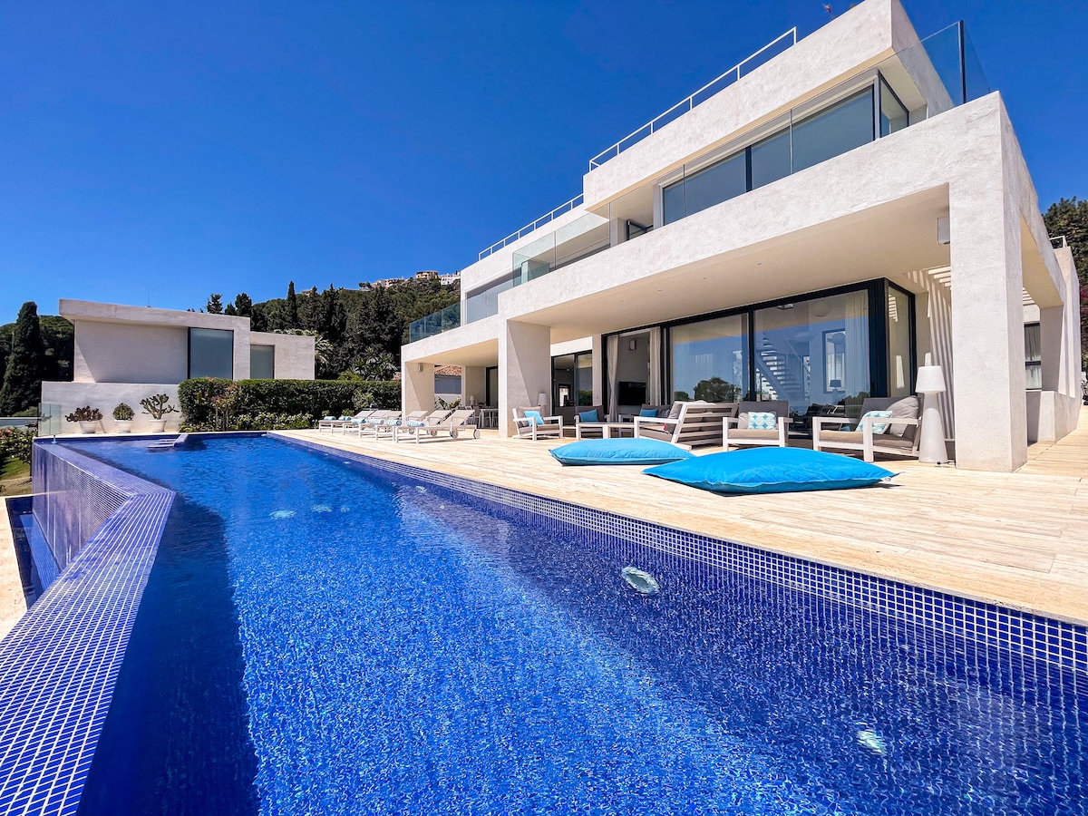 An expansive, modern villa is presented alongside a shimmering infinity pool, which reflects the clear blue sky. Multiple sun loungers are arranged along the poolside, providing ample space for relaxation. The stylish outdoor area is complemented by a backdrop of lush greenery.