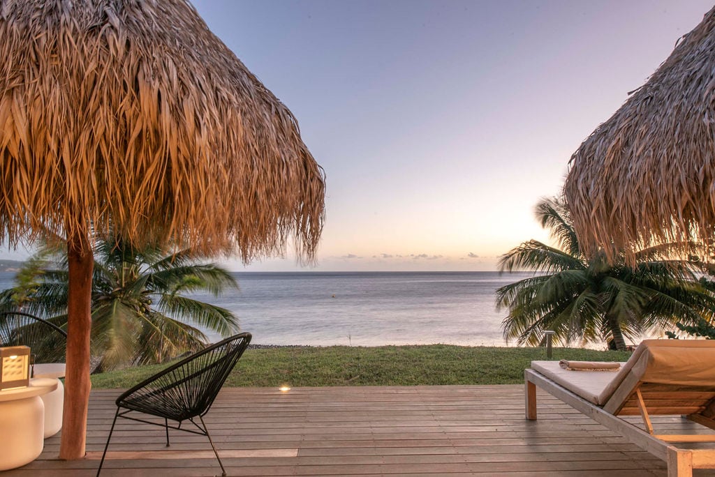 A spacious outdoor area features two thatched umbrellas providing shade over lounge chairs. The calm sea extends towards the horizon, framed by lush tropical plants. Soft lighting highlights the wooden patio, creating a serene and inviting space for relaxation.