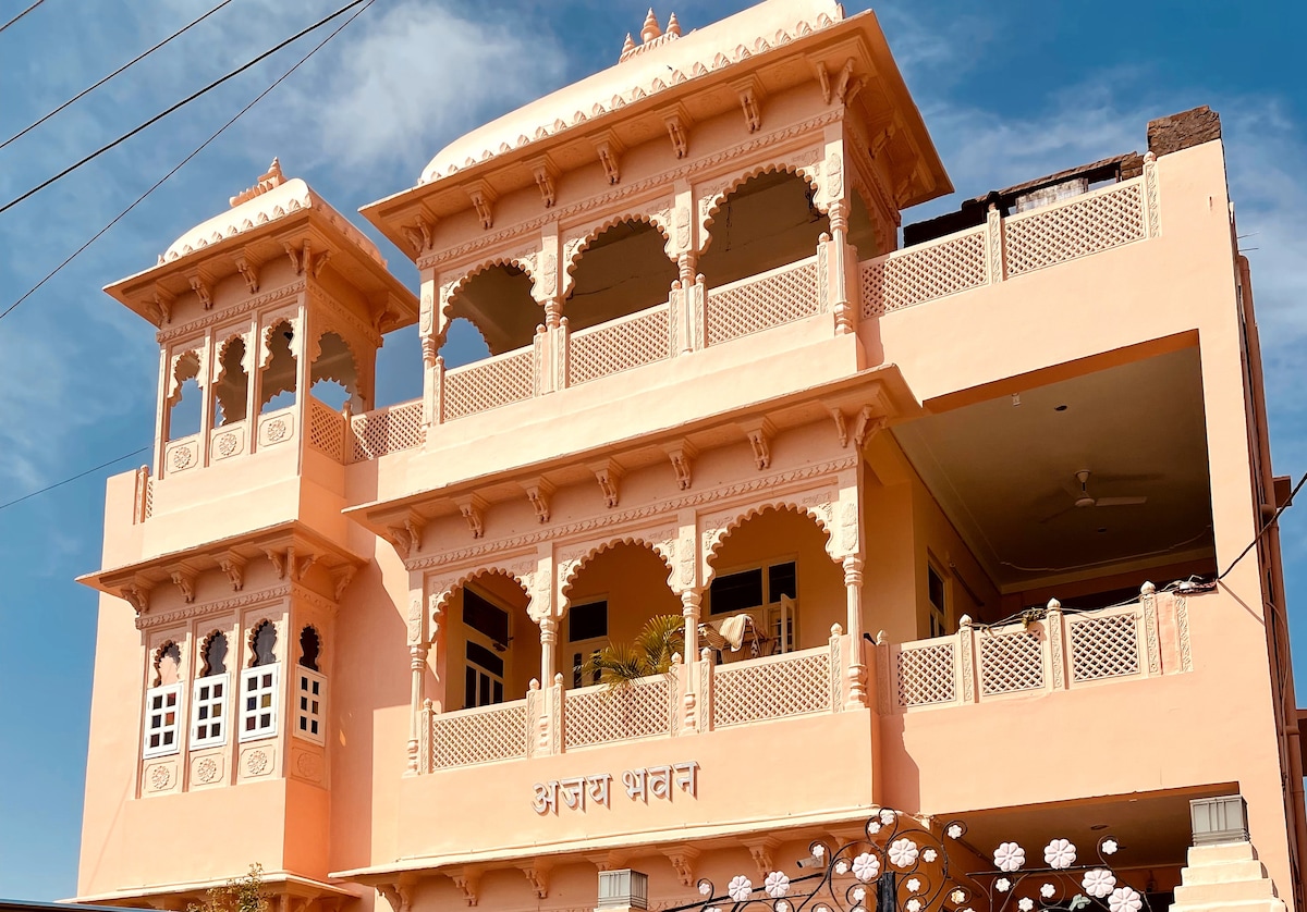 A charming two-story building features intricate architectural details, with a soft peach exterior and ornate balconies. Arched windows and decorative latticework enhance the façade, while a clear blue sky serves as a backdrop, emphasizing the beauty of the structure.