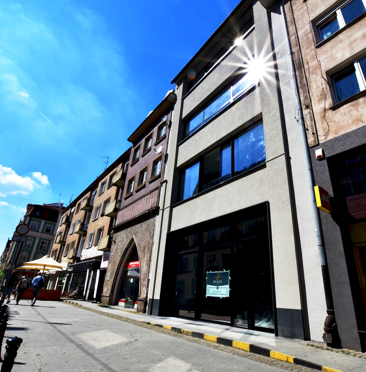 The building is viewed from a quaint street, showcasing a modern facade with large windows reflecting sunlight. The surrounding area features historical architecture, while outdoor seating is evident at nearby cafes. The sky is clear with a bright blue hue.