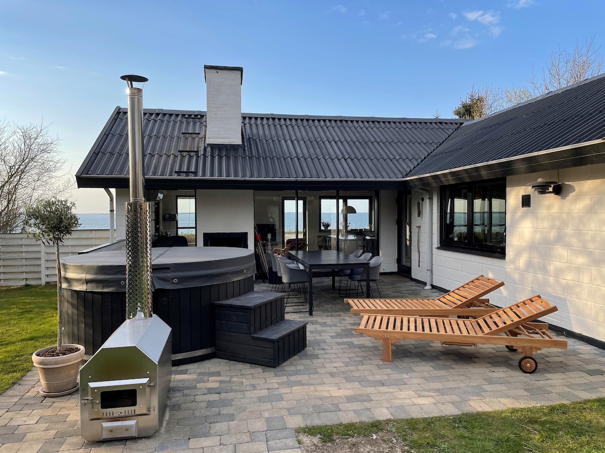 An outdoor area features a spacious wooden deck with lounge chairs and a hot tub. The hot tub, equipped with an external heater, is positioned near large glass doors that provide views of the waterfront. Lush green grass surrounds the area, enhancing the peaceful setting.