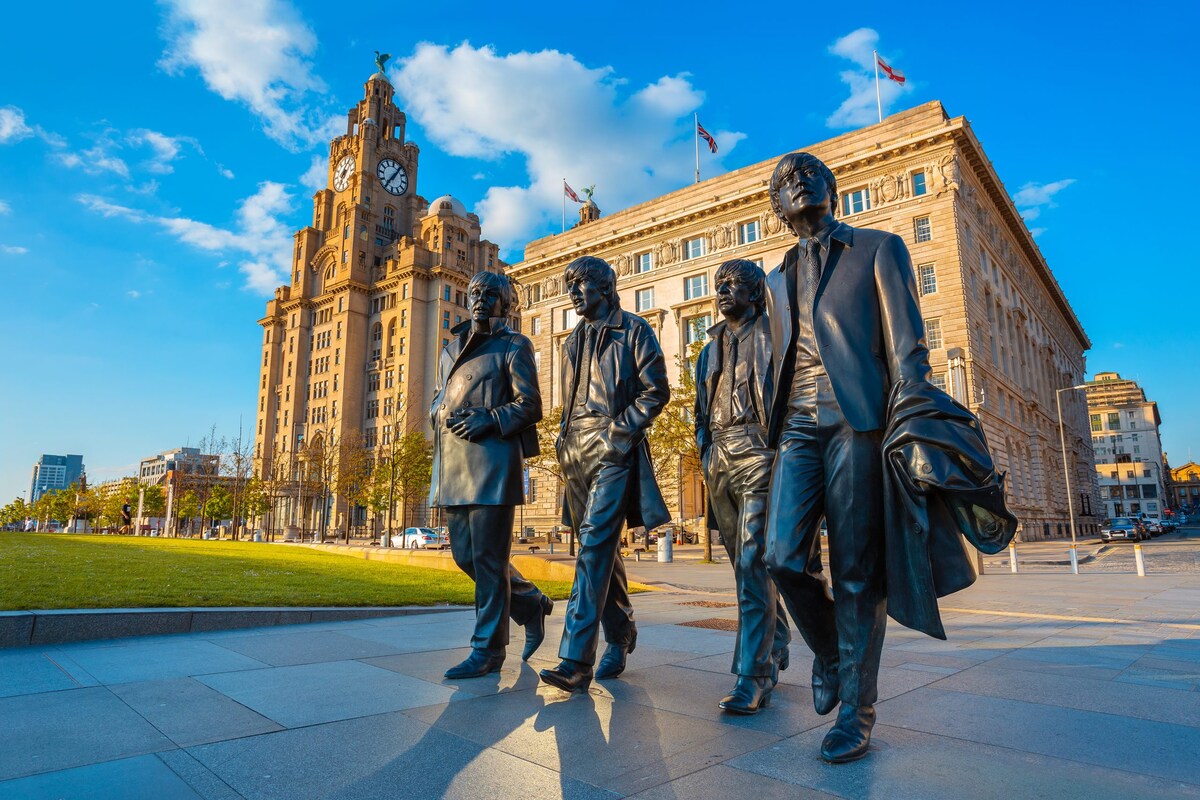Bronze statues of four figures are seen strolling together, set against the backdrop of an impressive historical building with a clock tower. The scene captures a vibrant sky with scattered clouds while a grassy area provides additional contrast in the foreground.