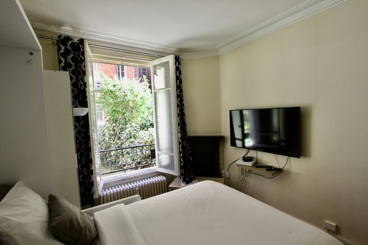 A bright room features large windows with black and white patterned curtains, allowing natural light to enter. A minimalist bed is positioned nearby, with a wall-mounted television visible opposite. A radiator is situated under the window, complementing the earthy tones of the decor.