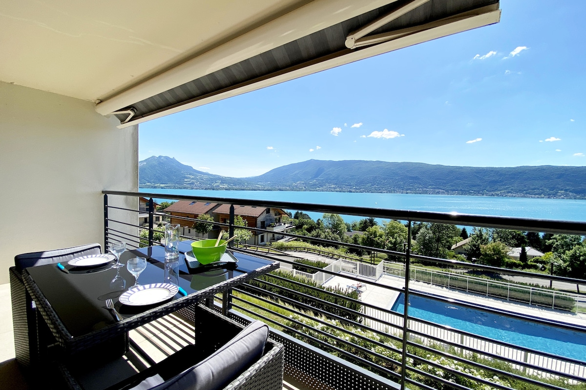 A private balcony is displayed, featuring outdoor furniture with a dining setup. A lake and mountain view is visible in the background, framed by clear blue skies. The nearby swimming pool can be seen below, enhancing the outdoor space.