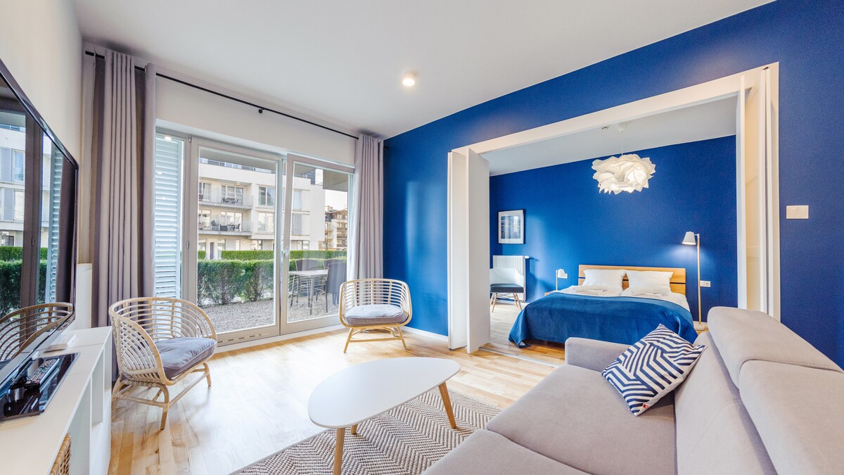 A spacious living area features a light-colored sofa and a round coffee table, set against a deep blue accent wall. Two rattan chairs provide additional seating, and large sliding glass doors lead to a courtyard. A partially visible bedroom includes a bed and an artistic light fixture.