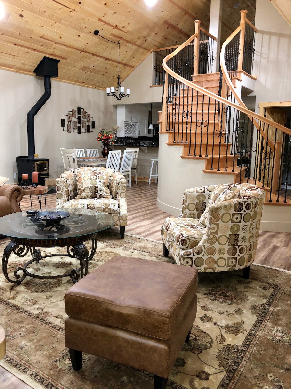 The living area features two patterned chairs surrounding a glass-top coffee table on a decorative rug. A staircase leads to the upper level, framed by wrought iron railings. A modern wood stove is positioned against the wall, while the kitchen is visible in the background.