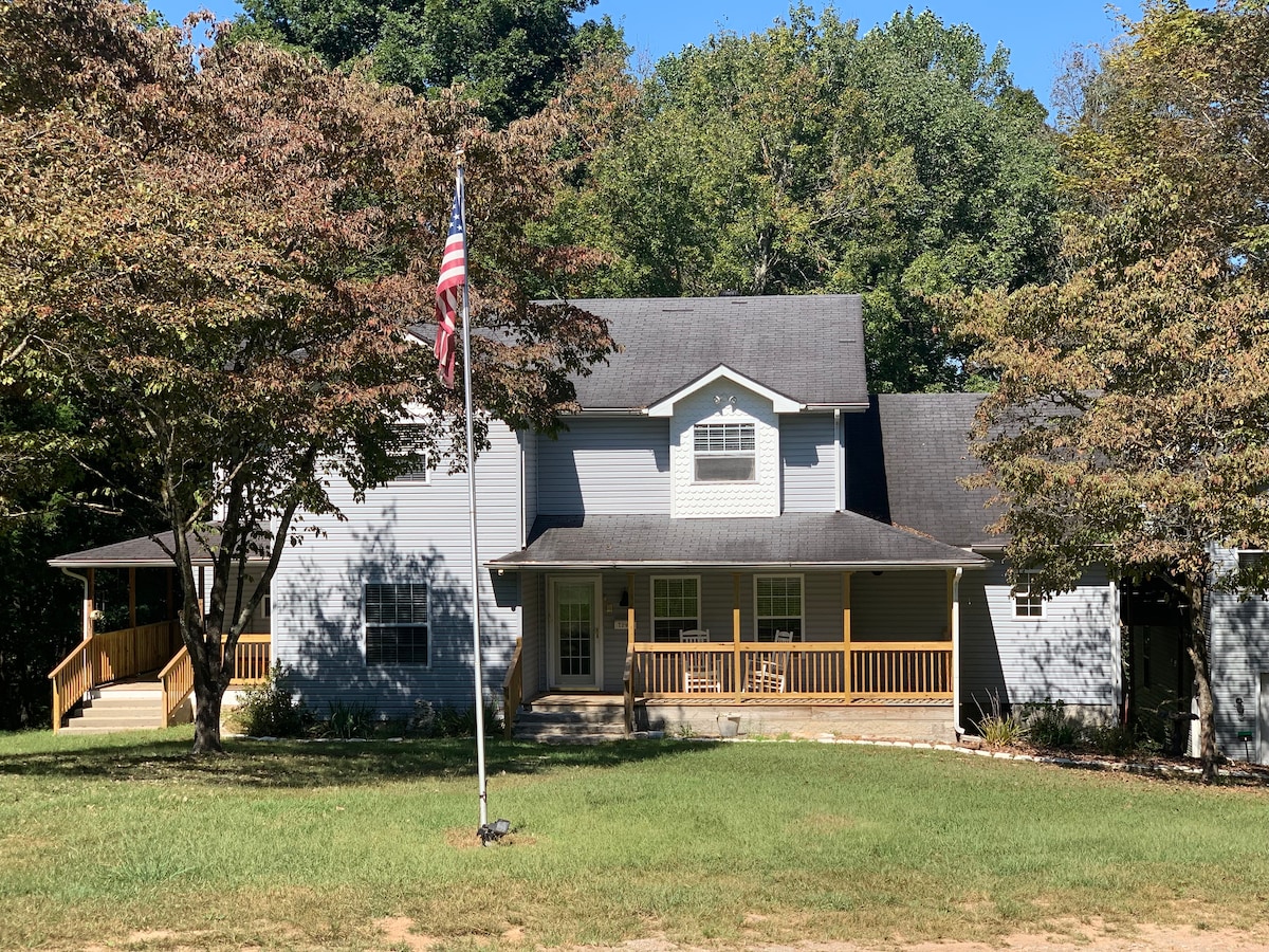 The exterior of a spacious grey house is framed by lush greenery, featuring a welcoming front porch with seating. A flagpole stands in the foreground, displaying the American flag. The well-maintained lawn enhances the serene atmosphere of the property.