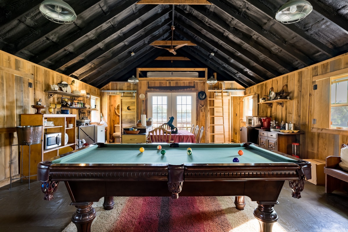 A spacious loft interior features a pool table centrally located, surrounded by rustic wooden walls. A kitchenette can be seen in the background, along with a dining area furnished with a round table and chairs. Natural light streams in through large windows.