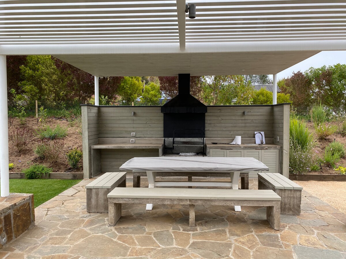 An outdoor dining area features a large stone table surrounded by matching benches. A built-in grill is positioned against a wooden backdrop. Lush greenery and landscaping can be seen in the background, adding to the serene outdoor setting.