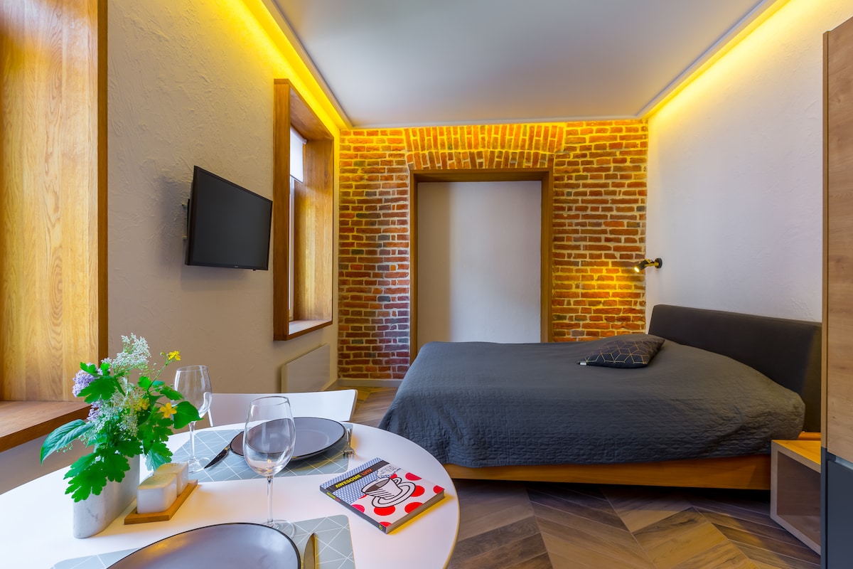 A modern studio apartment is featured with a spacious bed in the background and a dining table set for two in the foreground. Exposed brick walls and ambient lighting create a cozy atmosphere. A wall-mounted television is visible, enhancing the functional design of the space.