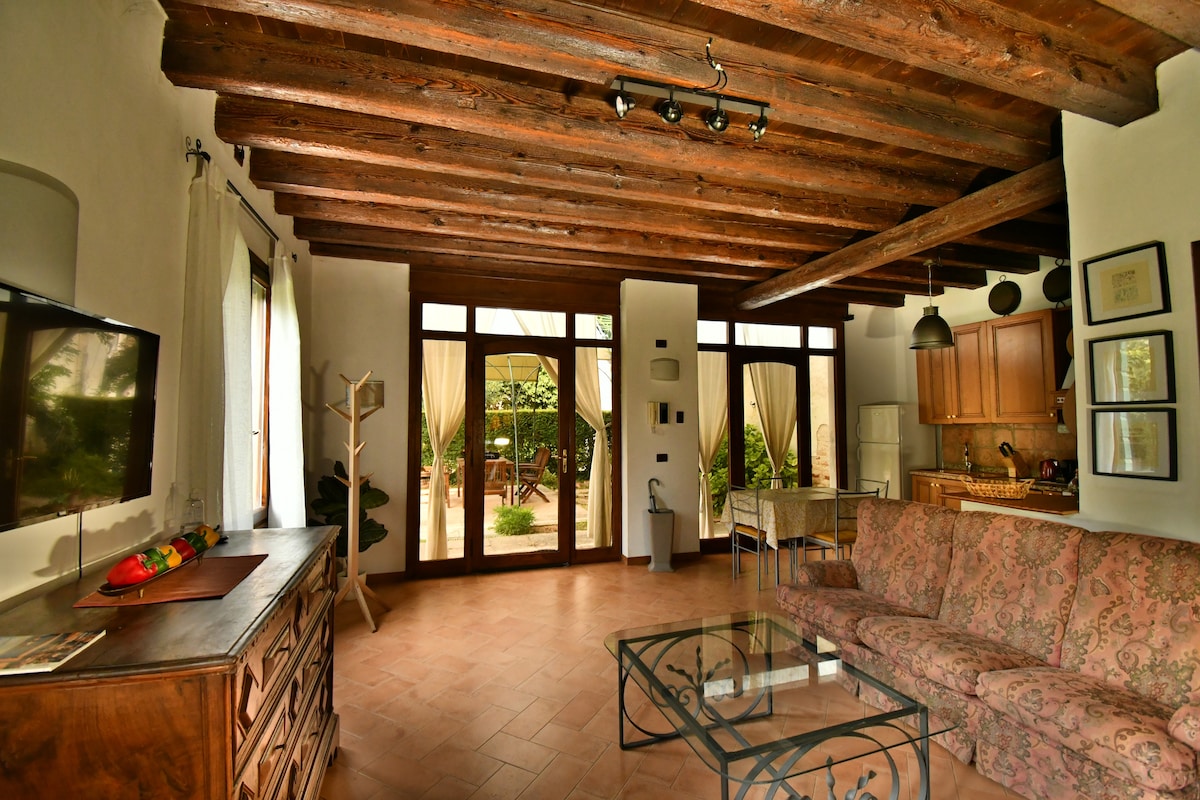 The living area features wooden beamed ceilings and large glass doors that provide a view of the private patio. A comfortable sofa and a glass coffee table are positioned in the space, while a wooden cabinet and dining area are also visible.