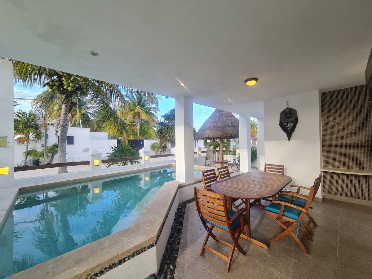 An outdoor seating area is showcased, featuring a wooden table surrounded by six chairs with blue cushions. A pool is visible nearby, framed by tropical plants and white walls. The space is well-lit, creating an inviting environment for relaxation.