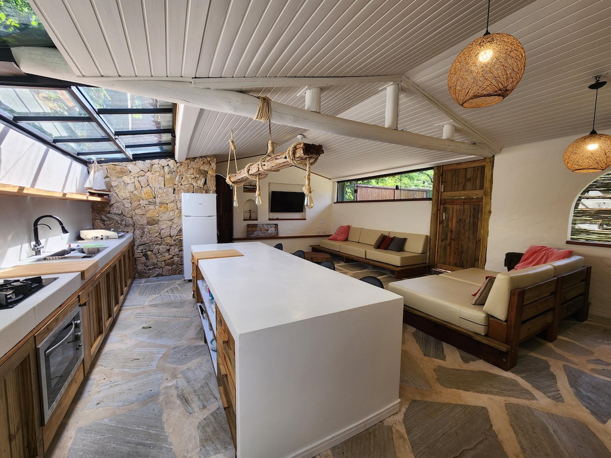 An open-plan kitchen and living area are showcased, featuring a well-equipped kitchen with modern appliances. Natural light streams in through large windows. Comfortable seating is arranged around a central island, with a stone wall adding texture to the space.