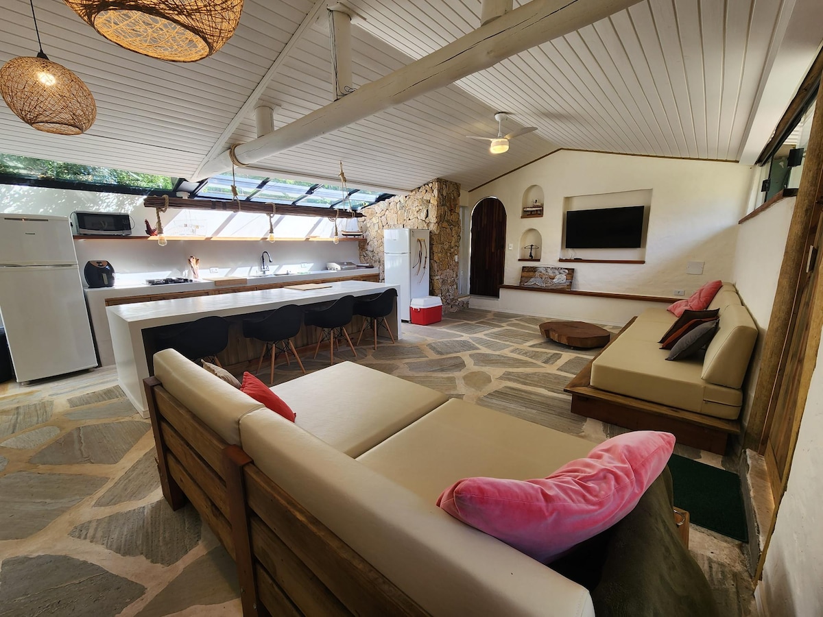 An open-concept living space features integrated kitchen and lounge areas. Natural stone flooring is combined with wooden seating and colorful cushions. A large window provides ample light, while a wall-mounted television is positioned for comfortable viewing. Decorative elements add charm to the neutral design.