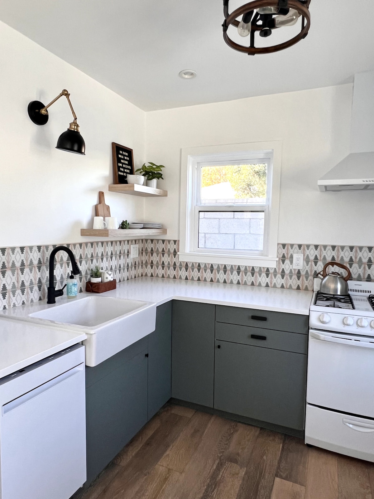 A modern kitchen features a spacious white countertop and a farmhouse-style sink. Stylish gray cabinetry and patterned tile backsplash add character. A window allows natural light, while a sleek stove and kettle complete the well-appointed cooking area.