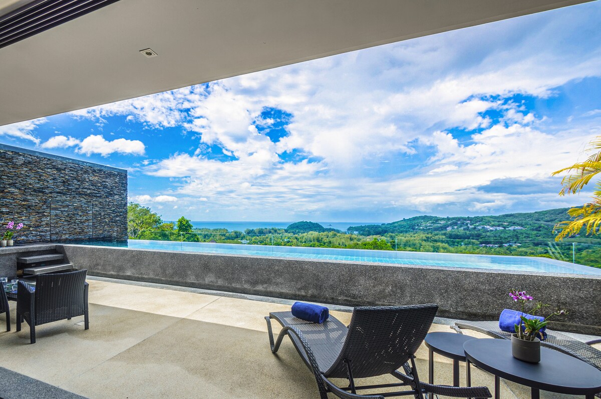 An outdoor terrace features lounge chairs positioned for relaxation, overlooking a stunning panoramic view of the Andaman Sea and lush greenery. An infinity pool reflects the sky's colors, enhancing the serene ambiance of the private retreat.