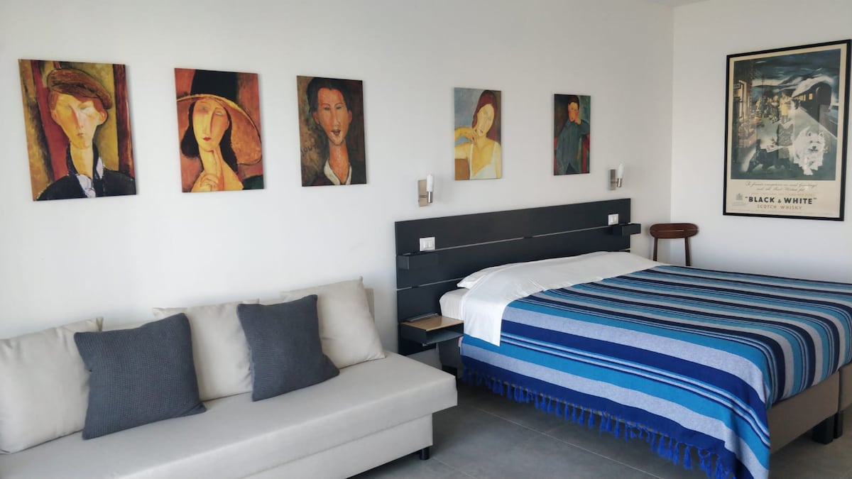 A modern bedroom features a spacious bed with a blue and white striped blanket. A light-colored sofa with throw pillows sits nearby. Abstract art pieces adorn the walls, complemented by soft lighting from wall sconces.