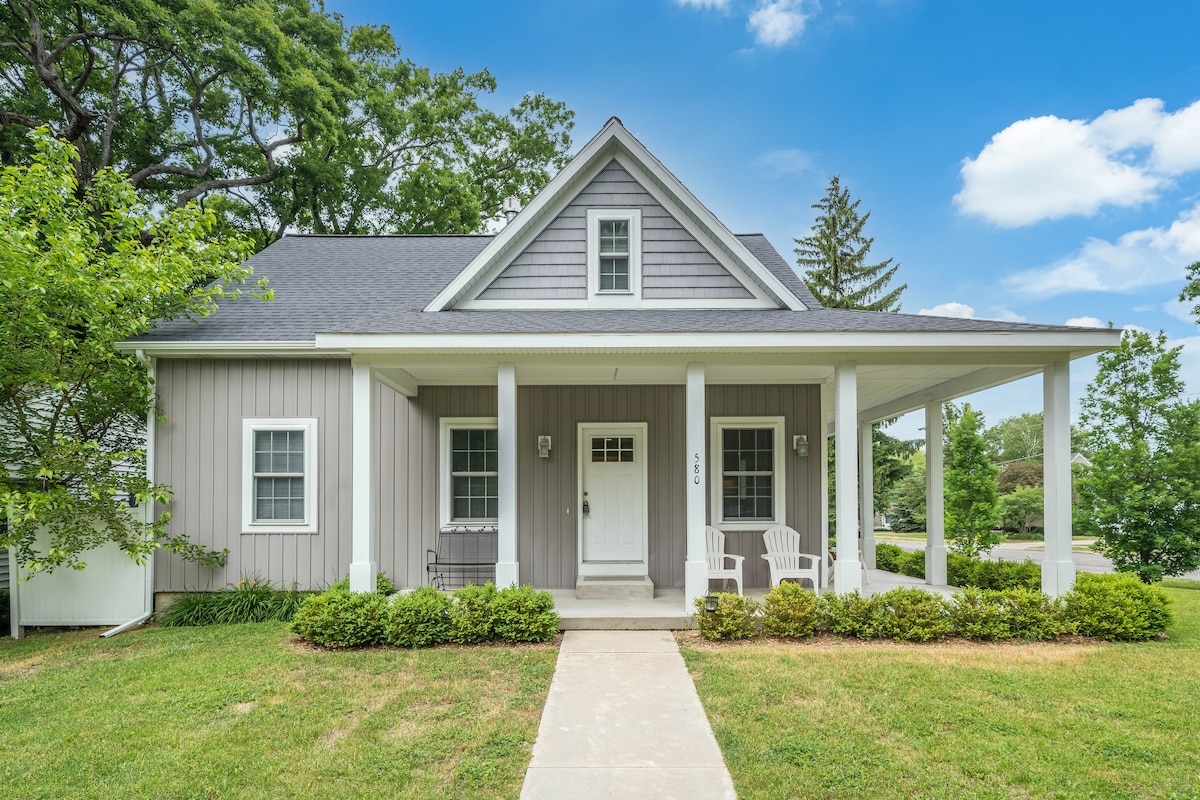 A charming exterior of the cottage features a covered porch with two white adirondack chairs. The house is painted in soft gray tones with white trim and a triangular gable above the entryway. Lush green grass and shrubs frame the pathway leading to the door.
