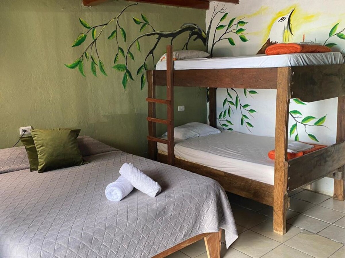A cozy sleeping area features a spacious bed draped in soft linens beside a wooden bunk bed. Playful mural artwork of leaves and a bird adorns the wall, creating a vibrant, nature-inspired ambiance. Light-colored tiled floors contribute to the room's open feel.