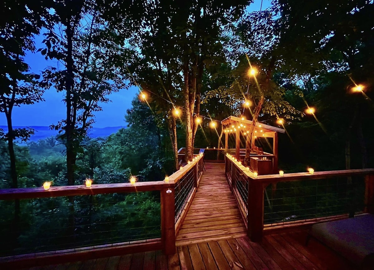 A wooden pathway adorned with string lights leads to a cozy seating area among trees. The evening sky is softly illuminated, enhancing the natural beauty of the surrounding woodland. The distant view displays rolling hills, creating a serene atmosphere.