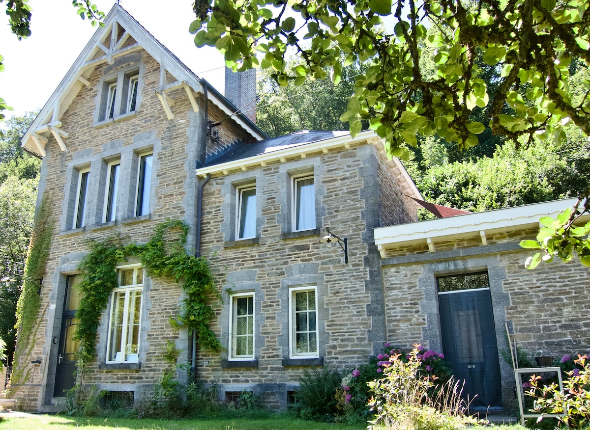 A stone house with a historic charm is enveloped by greenery. The structure has large windows and a welcoming porch awning. Climbing plants adorn the facade, while colorful flowers are seen in the front garden, contributing to the serene outdoor setting.