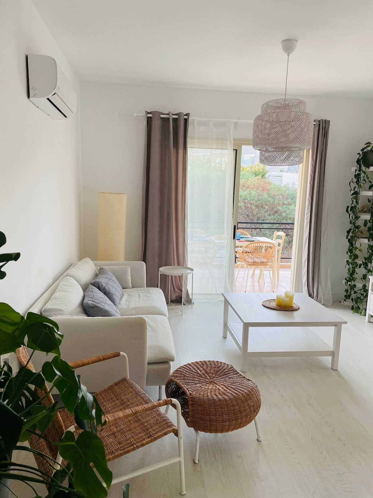 The living area is characterized by a light-colored sofa and a small round table. Natural light enters through large glass doors, leading to the balcony. A stylish pendant lamp hangs above, while greenery and soft fabric curtains enhance the space's comfort.