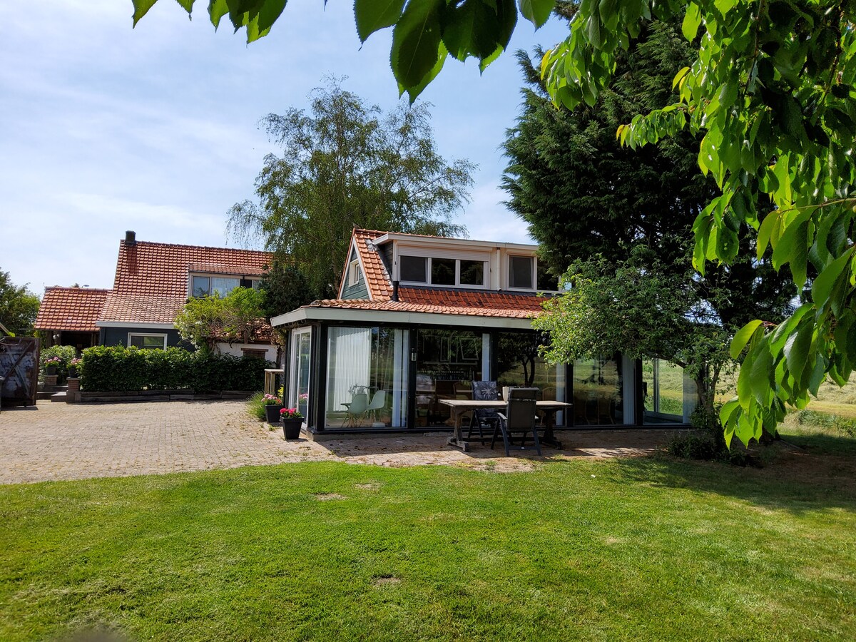 A welcoming house features a red-tiled roof and a glass-enclosed terrace, surrounded by green grass. Large windows provide views of the outdoor space, which includes trees and plants. The area is spacious, inviting relaxation and enjoyment of the natural surroundings.