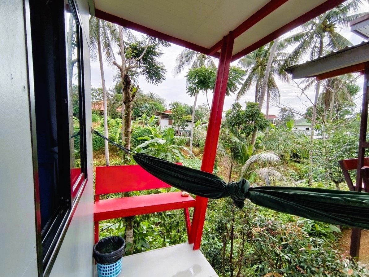 A balcony area presents a suspended hammock, inviting relaxation amidst lush greenery. Surrounding palm trees and plants create a natural ambiance, with a small bench positioned nearby. The exposed red beams of the bungalow frame the view, contributing to a serene outdoor experience.