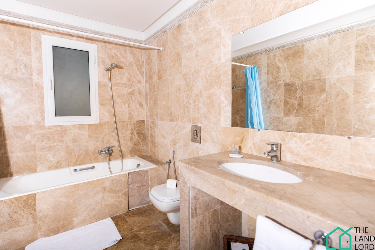 The bathroom features marble walls and flooring, providing a spacious feel. A bathtub is positioned alongside a separate shower. A modern toilet and a vanity with a round sink are present, complemented by a mirror above and a towel laid neatly on the floor.