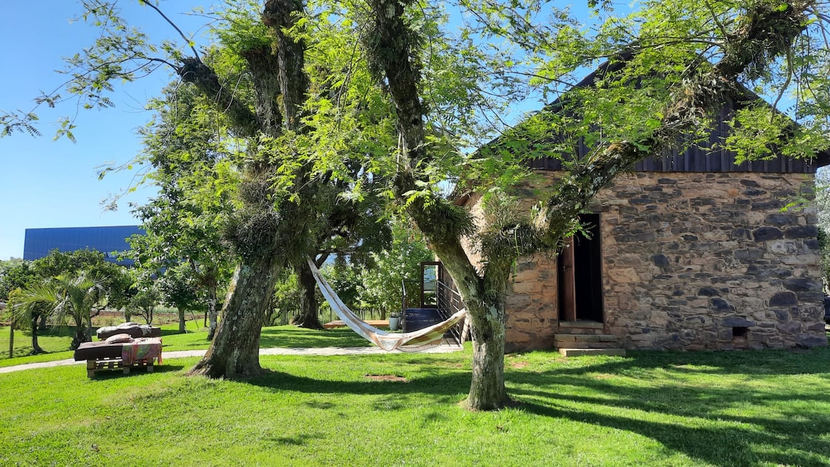 A rustic stone house is framed by large trees with abundant green foliage. A hammock is suspended between two trees, inviting relaxation. The surrounding lawn is well-kept, with hints of additional landscaping elements subtly visible in the background.