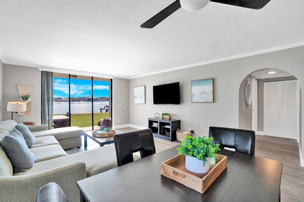 A bright living area features a comfortable sectional sofa and a coffee table, with large glass doors revealing scenic waterfront views. A dining set with dark chairs and a decorative tray adds functionality. Light is softly diffused through the space, creating a welcoming ambiance.