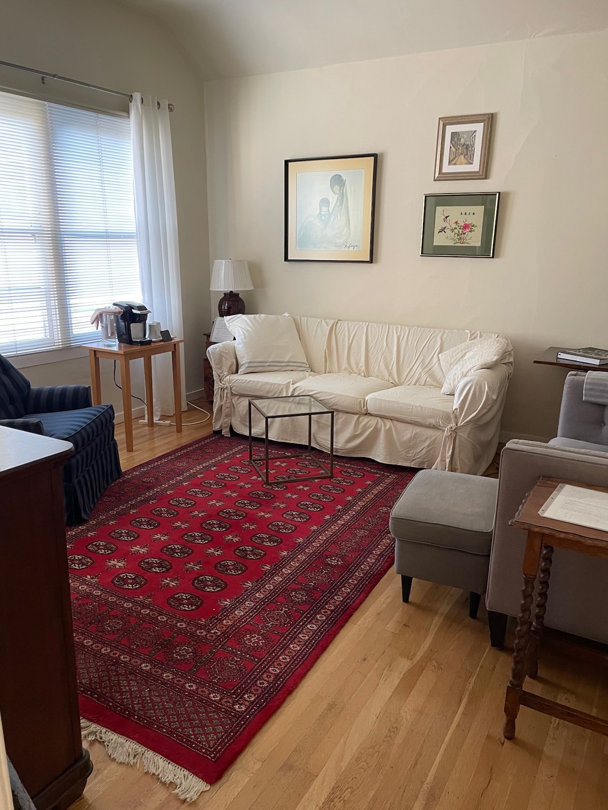 A cozy living area showcases a light-colored sofa with a contrasting decorative pillow. A low coffee table sits atop a patterned area rug. Natural light filters through multiple window panes, illuminating framed artworks and a small coffee station in the corner.