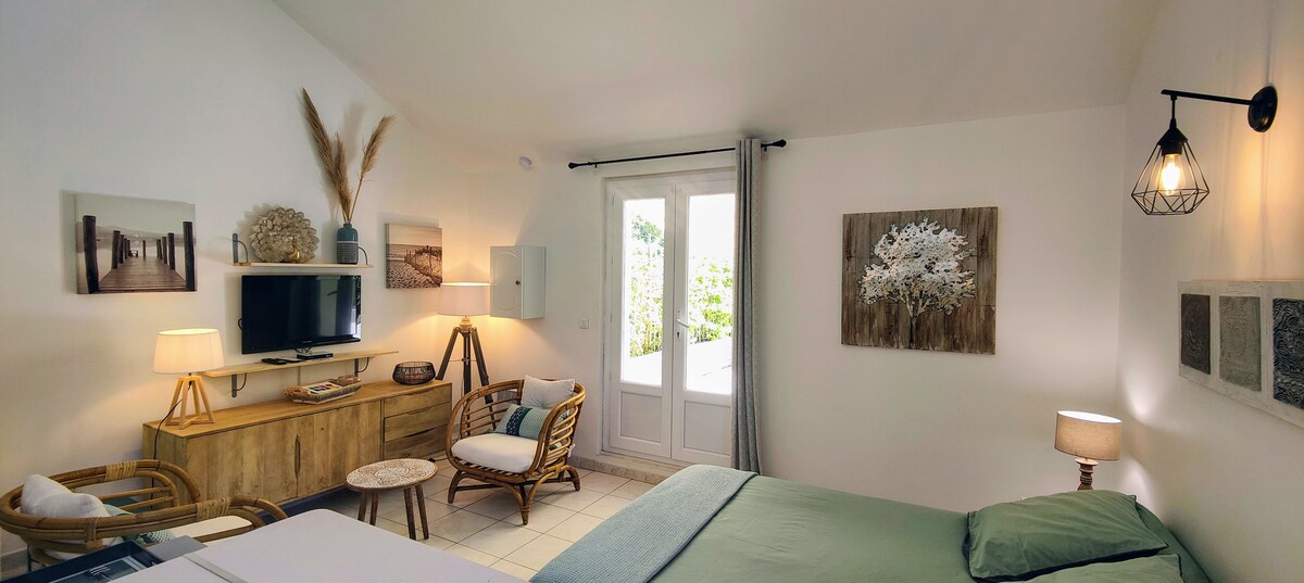 A well-appointed room features natural light from large windows and a soothing color palette. A comfortable bed is accompanied by wicker furniture, including a chair and a small table. Decorative wall art adds a touch of elegance to the tranquil space.
