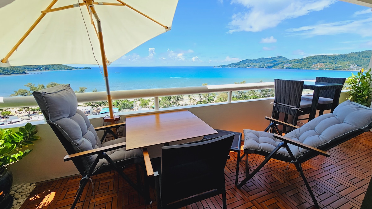 A west-facing balcony features comfortable seating, including two lounge chairs and a table. A large umbrella provides shade while a panoramic view captures the shimmering blue waters of Patong Beach and lush hills in the background.