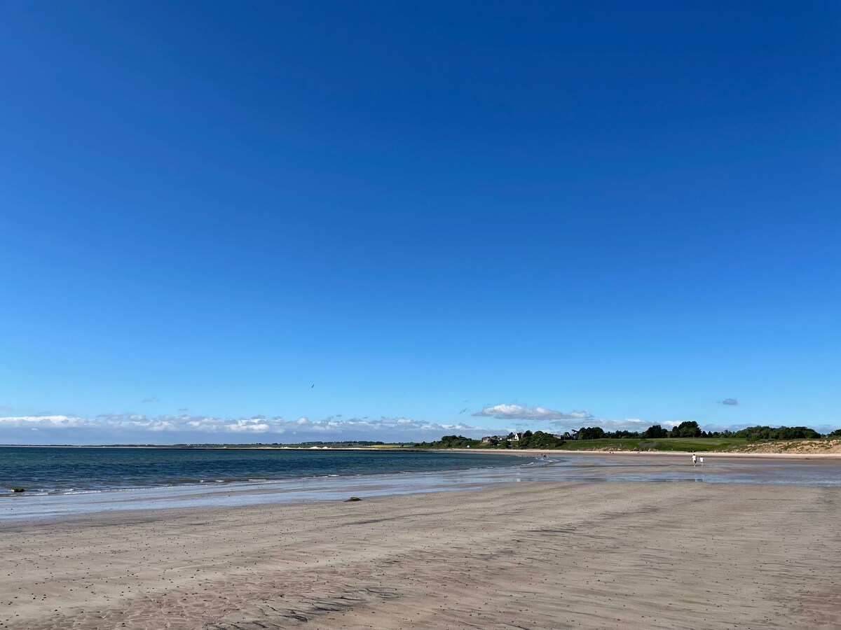 A wide sandy beach stretches beneath a clear blue sky, with gentle waves lapping at the shore. The tranquil scene is complemented by distant grassy dunes and patchy clouds, creating a serene coastal atmosphere.