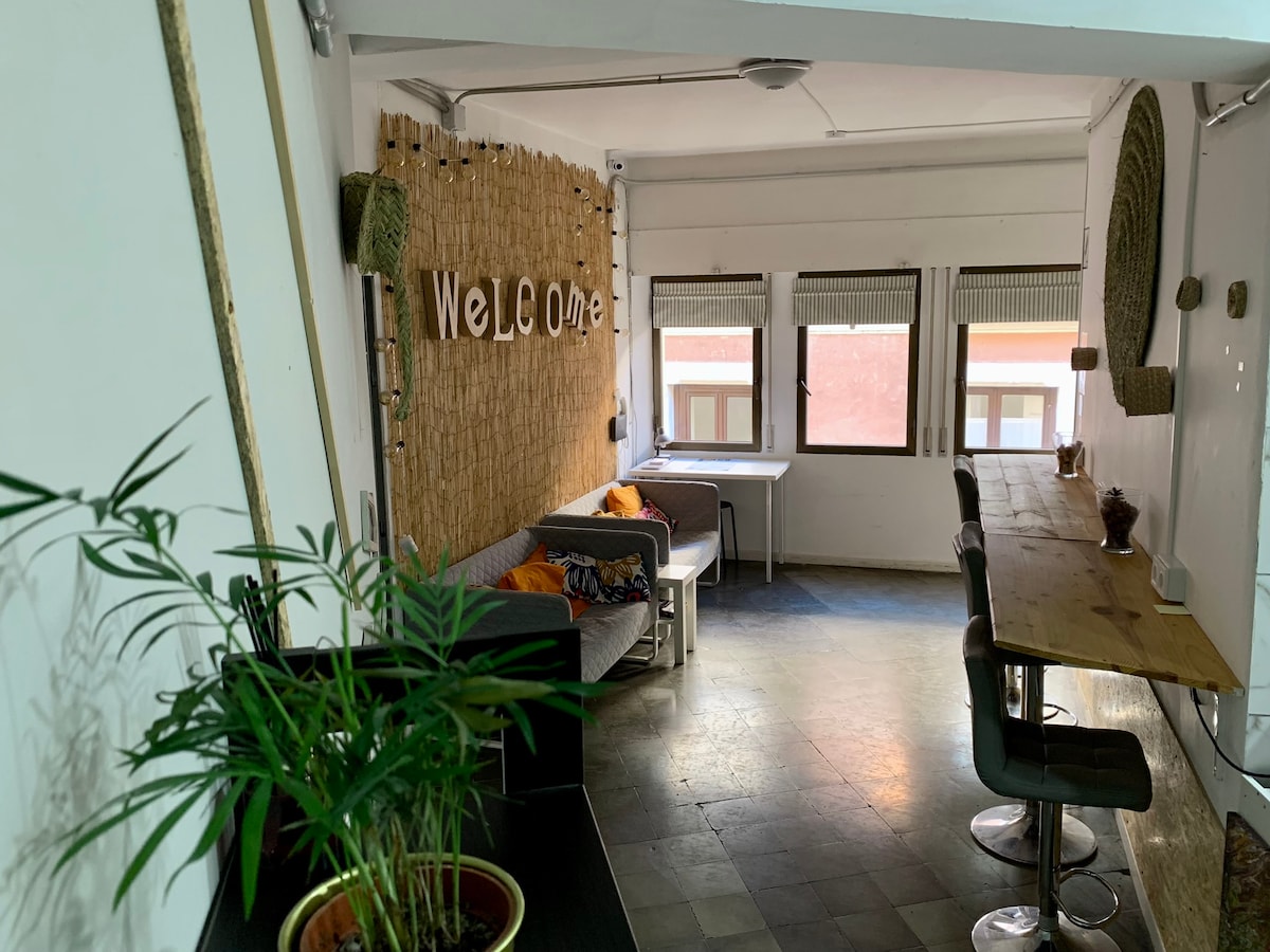 A bright common area features a large dining table with high stools, surrounded by natural light from multiple windows. A welcoming sign decorates the wall, and cozy seating is provided with colorful cushions. Indoor plants add a touch of greenery to the space.