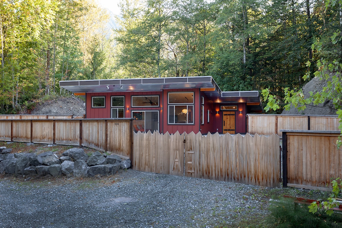 A modern cabin features a combination of red and wood finishes, surrounded by a natural landscape. A low wooden fence spans the front, and smooth gray stones are visible in the foreground. Windows are framed with dark accents, enhancing the building's contemporary design.