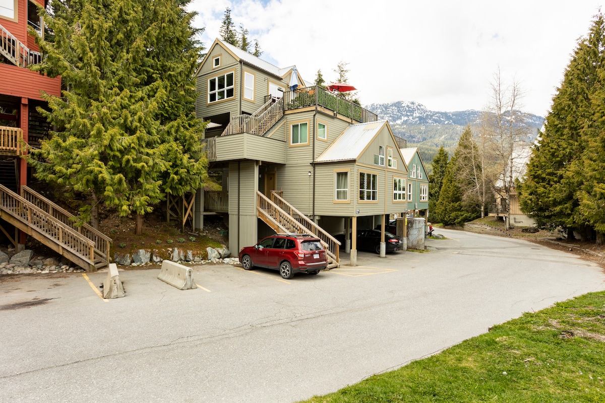 A modern townhouse is pictured, nestled among tall coniferous trees. The exterior features green and grey tones, with large windows that reflect the surrounding landscape. A parking area stands in front, accommodating several vehicles, while a peaceful street leads away from the property.