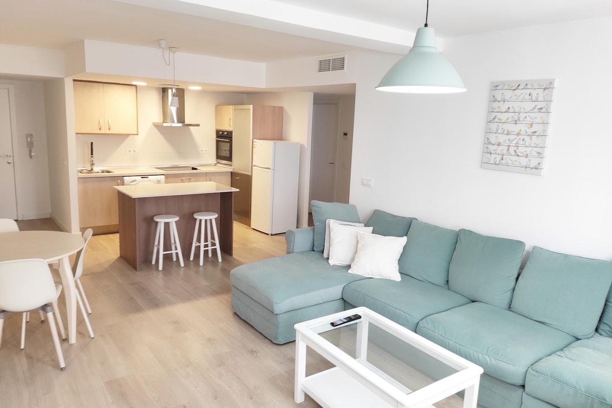 A spacious living area features a modern light blue sectional sofa with decorative cushions. A glass coffee table sits in front, and a dining table with white chairs is visible. The open kitchen has light wood cabinets, a stovetop, and a refrigerator.