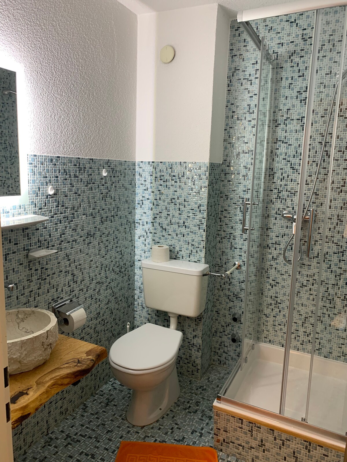 The compact bathroom features artistic blue mosaic tiles covering the walls, with a glass shower enclosure visible. A unique stone sink is mounted on a wooden countertop, while a toilet is situated against a wall for efficient use of space. Natural light is provided by a small window.