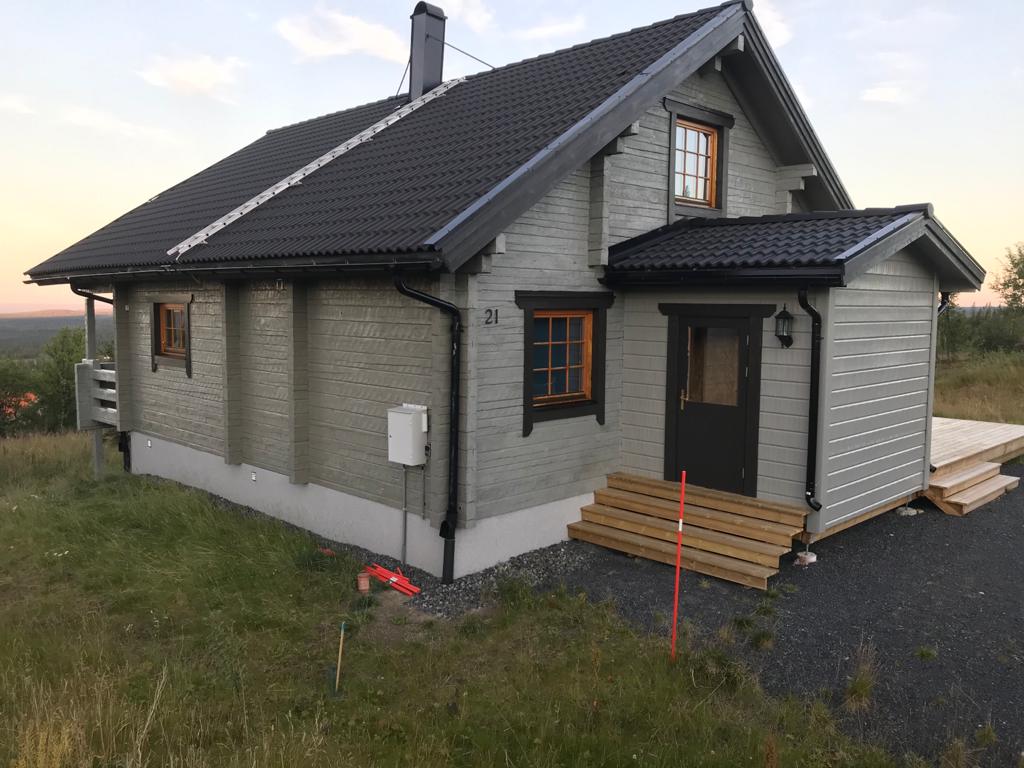 The exterior of a wooden cabin showcases a light grey facade complemented by dark wooden accents and windows. A wooden deck is visible at the entrance, surrounded by grassy areas. The setting is tranquil, enhanced by a soft evening light illuminating the structure.