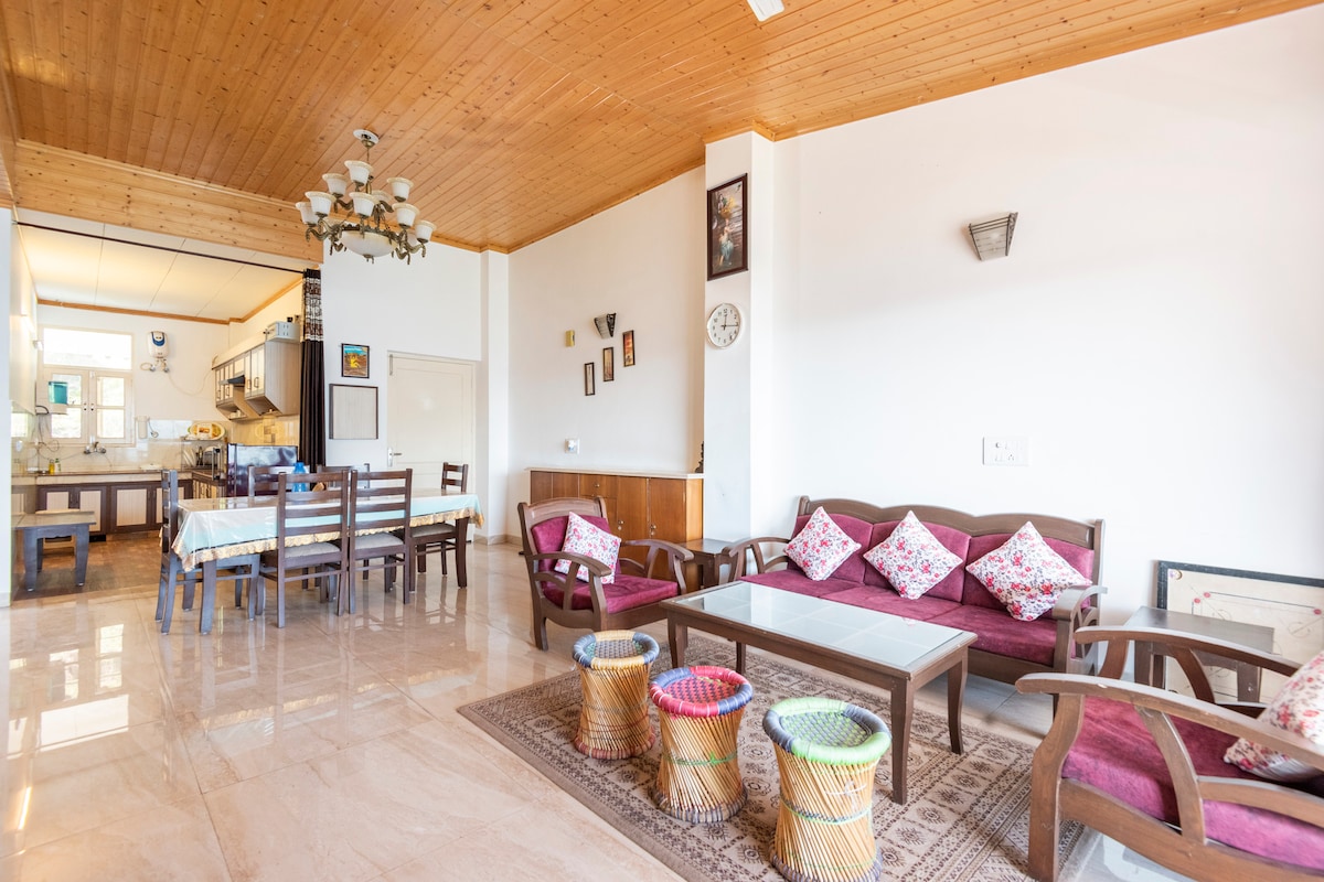 A spacious living area features wooden ceiling panels and elegant light fixtures. A comfortable seating arrangement with colorful cushions is complemented by a light-colored coffee table. A distinct dining area with wooden chairs is visible in the background, along with an open kitchen space.
