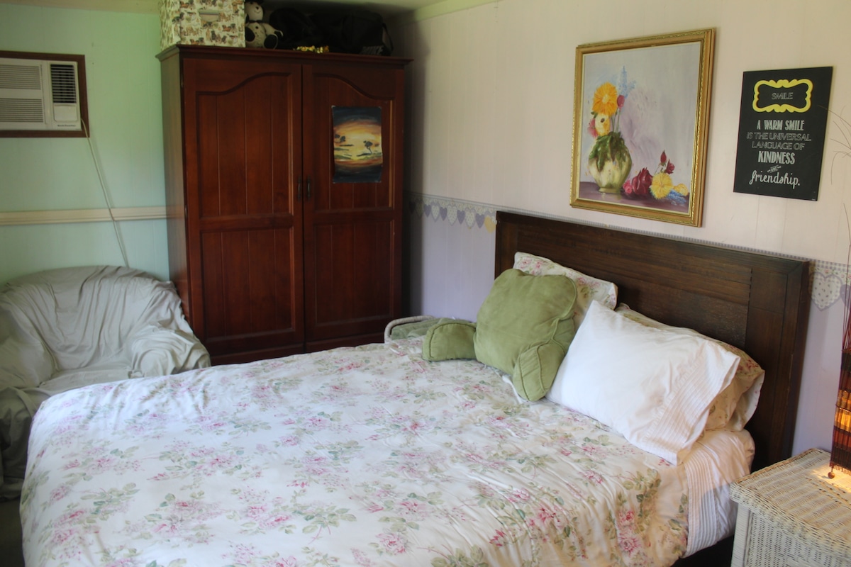 A cozy bedroom features a spacious bed adorned with floral bedding, alongside a light-colored armchair. A wooden wardrobe stands against the wall, and an air conditioning unit is positioned above the bed. A piece of framed artwork and a decorative sign add a touch of charm.