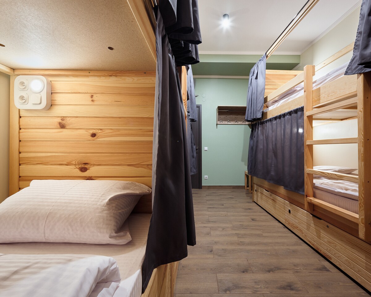 A shared dormitory is presented with wooden bunk beds, framed by simple grey curtains for privacy. Each bed features a comfortable mattress and pillows. Warm wood accents contribute to a cozy atmosphere, and the floor is covered with neutral-toned planks.