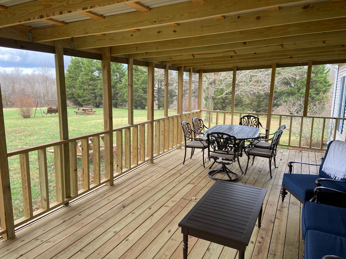 A covered porch is presented with wooden flooring and a rustic railing. A dining table with six chairs is positioned centrally, accompanied by a seating area featuring two blue chairs and a small black table. Lush greenery surrounds the space, enhancing the outdoor experience.