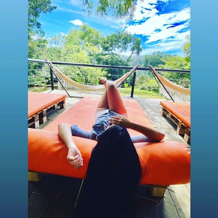 A woman reclines on an orange lounge chair, surrounded by a lush green landscape. Two hammocks are positioned nearby, framing the serene view of trees and a blue sky above. The setting offers a peaceful retreat in nature.