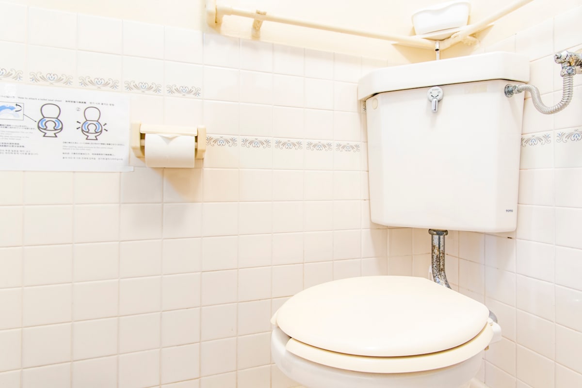 The bathroom is presented with a clean and functional toilet, featuring a decorative wall with light-colored tiles. A toilet paper holder is mounted nearby, ensuring convenient access. Instructions are visible on the wall, contributing to user-friendly functionality.
