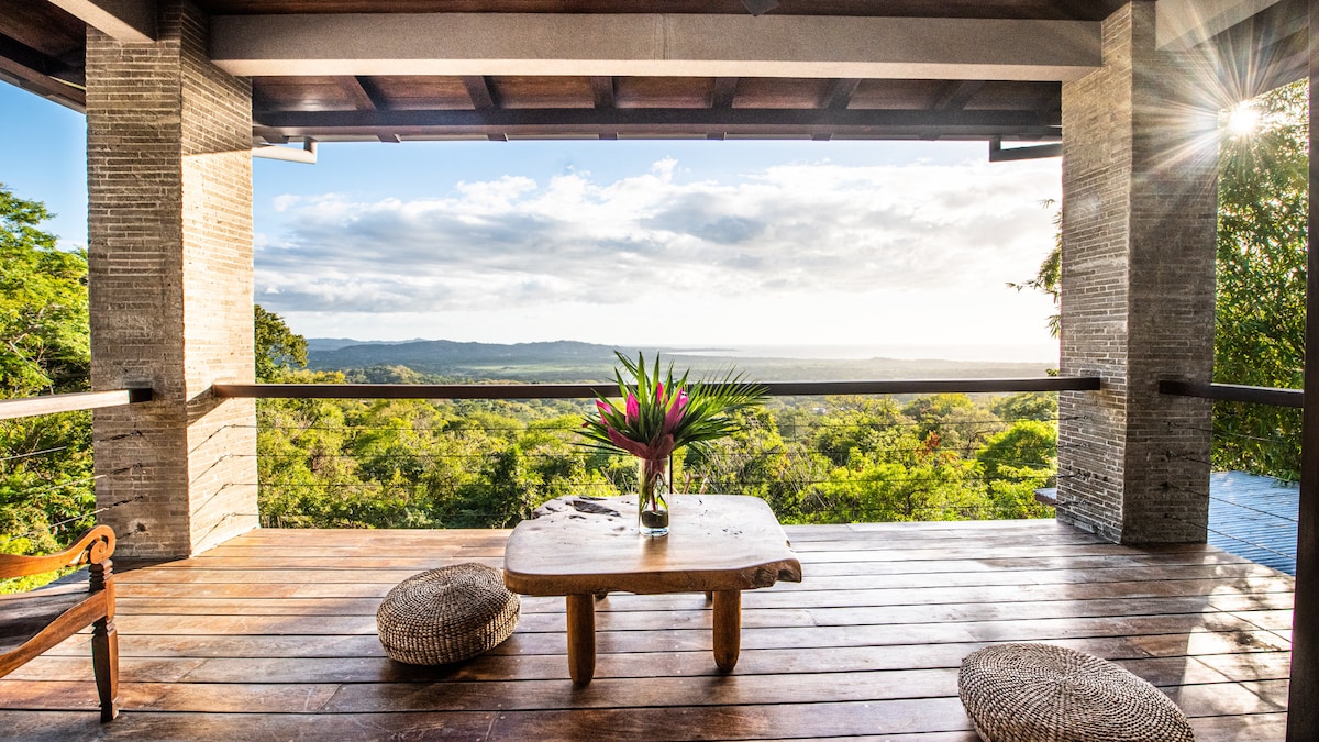A spacious outdoor terrace features a wooden table adorned with a tropical plant centerpiece. Comfortable round seating surrounds the table, and expansive openings reveal stunning views of the lush green landscape and sky, inviting a tranquil atmosphere.